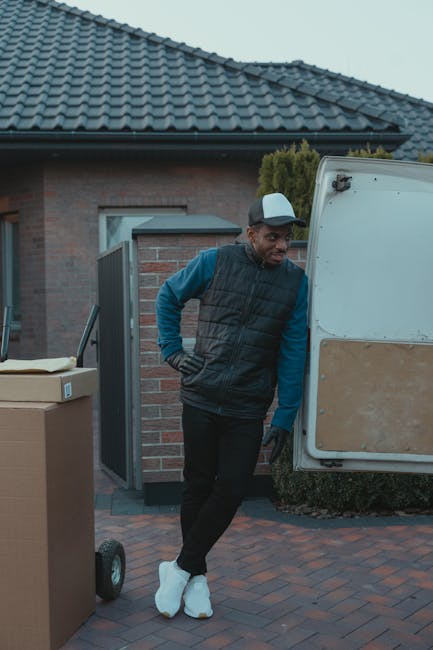 A man dressed in a blue long-sleeved shirt, black padded vest, and black trousers, wearing white sneakers and a grey cap, standing outside a residential property next to a white moving van with a wood