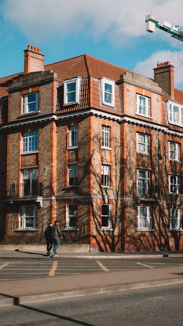 A row of multi-storey terraced houses with brick facades, some featuring bay windows and slate roofs, lining a busy street in Carshalton. The street includes a shopfront with a blue and white sign, pe