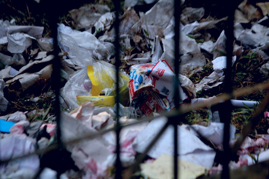 A view through a black metal fence showing trash and litter on the ground outdoors, including crumpled paper, plastic bags, and food packaging. The debris is scattered on a patch of grass and soil, with some leaves mixed in. The image depicts neglected waste accumulation, separate from the typical packing materials used during home removals or furniture transport. This scene relates to waste disposal and disposal rules, which are relevant to house removals services such as those offered by Man with Van Carshalton, especially in the context of local regulations like those in Sutton Council. The environment is unkempt and shows no furniture or packing supplies, contrasting with professional relocation processes involving safe handling, packing, and transport of belongings.