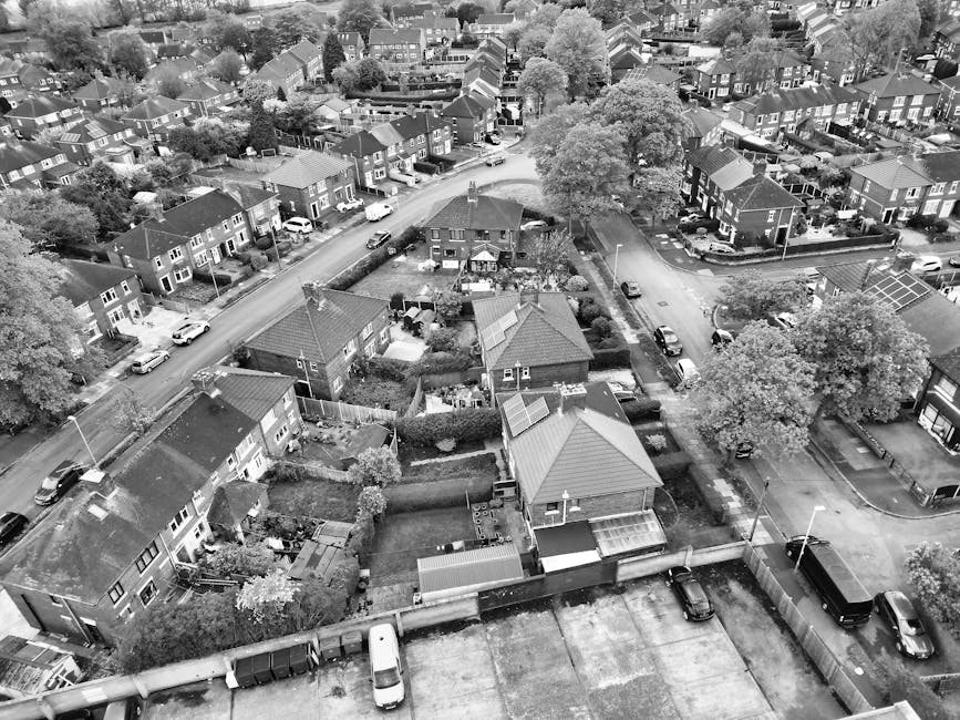 An aerial view of a residential neighbourhood showing rows of houses with pitched roofs, some with solar panels, and well-maintained gardens separated by hedges and fences. The scene includes parked cars along the streets and driveways, with a larger black van parked on the pavement near a house. In the foreground, roof sections and a flat rooftop area are visible, with some boxes and items likely prepared for a house move. The streets are quiet, and the overall environment reflects a typical suburban area suitable for home relocation services. This image illustrates the transportation and logistical considerations involved in furniture transport and packing during a home removal, with Man with Van Carshalton expertly managing these activities in a residential setting.