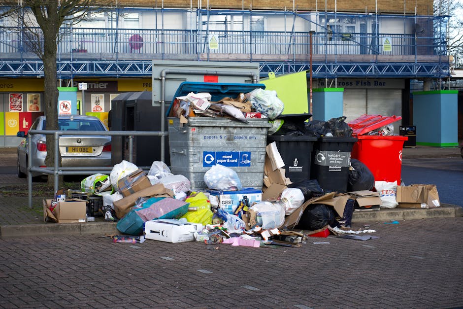 A view through a black metal fence showing trash and litter on the ground outdoors, including crumpled paper, plastic bags, and food packaging. The debris is scattered on a patch of grass and soil, with some leaves mixed in. The image depicts neglected waste accumulation, separate from the typical packing materials used during home removals or furniture transport. This scene relates to waste disposal and disposal rules, which are relevant to house removals services such as those offered by Man with Van Carshalton, especially in the context of local regulations like those in Sutton Council. The environment is unkempt and shows no furniture or packing supplies, contrasting with professional relocation processes involving safe handling, packing, and transport of belongings.