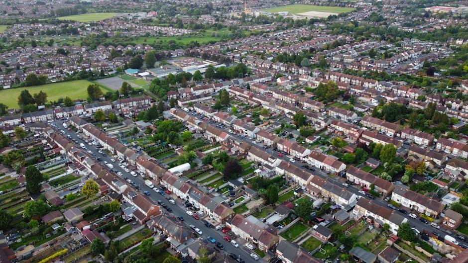 An aerial view of a residential neighbourhood in Carshalton, showing densely packed terraced houses with pitched roofs, small front gardens, and narrow streets lined with parked cars. Several houses have visible driveway areas, some with vehicles parked in them. The image captures a mix of greenery with trees and lawns interspersed among the houses, and open fields visible in the distance. The scene is well-lit with natural daylight, highlighting the urban layout and the proximity of homes to additional green spaces, which may present challenges for home relocation and furniture transport services. This setting illustrates a typical suburban environment where moving companies like Man with Van Carshalton assist with packing, loading, and transporting household items through narrow streets and limited access points, focusing on the logistics involved in relocating homes in difficult-to-access areas within SM5.