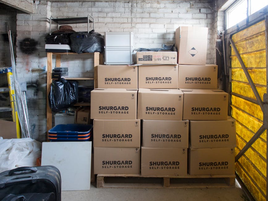 Inside a garage or storage unit with a brick wall background, stacked brown cardboard boxes labeled 'SHURGARD SELF-STORAGE' on a wooden pallet, arranged in a grid pattern. To the left, there are black plastic bags placed on a wooden shelving unit, and below them are smaller boxes and plastic containers. A metal ladder leans against the wall, and various packaging materials, including plastic wraps and packing boxes, are visible throughout the space. To the right, a partially open yellow vehicle door reveals a glimpse of a moving van, indicating the ongoing process of loading or unloading during a home relocation. The scene is illuminated by natural daylight, suggesting daytime activity, with the environment oriented towards preparing furniture, packed boxes, and belongings for transport. The setting reflects a typical moving or storage task managed by [COMPANY_NAME], focusing on packing, stacking, and preparing items for transportation in a household move as seen in the context of navigating difficult-to-access homes in SM5, Carshalton.