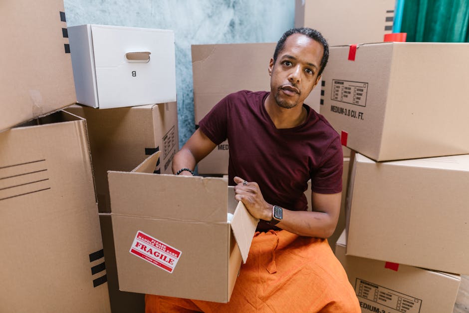 A man is kneeling among numerous cardboard boxes inside a room, likely during a home relocation, with some boxes stacked around him and others open, revealing packing materials. He is wearing an orange pair of trousers, a maroon T-shirt, and a wristwatch, and is looking directly at the camera. The boxes vary in size and are made of brown corrugated cardboard, some with red 'Fragile' labels and black printed barcode and description markings. A white cardboard box is placed on top of a stack in the background. The environment appears to be a moving or packing area within a house, with a textured blue-painted wall visible behind him. This scene illustrates the packing and loading process typically associated with professional removals, as performed by [COMPANY_NAME], emphasizing careful handling of household items before transport in a van or removal vehicle, and highlights the detailed organization involved in home furnishings transport and furniture moving.