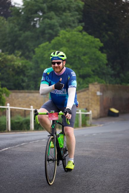 A man wearing a blue cycling jersey with white and green accents, black shorts, bright yellow cycling shoes, and a white helmet with blue and green stripes, is riding a black and green road bicycle on a paved street. He has sunglasses and a beard, and is smiling while giving a thumbs-up gesture. In the background, there is a stone wall and lush green trees, with an overcast sky overhead. This scene captures the man actively cycling outdoors, potentially as part of a home relocation or moving logistics process involving outdoor travel or transport, reflecting the services of Man with Van Carshalton. The image emphasizes outdoor mobility and active transportation, relevant to the theme of moving or logistics.