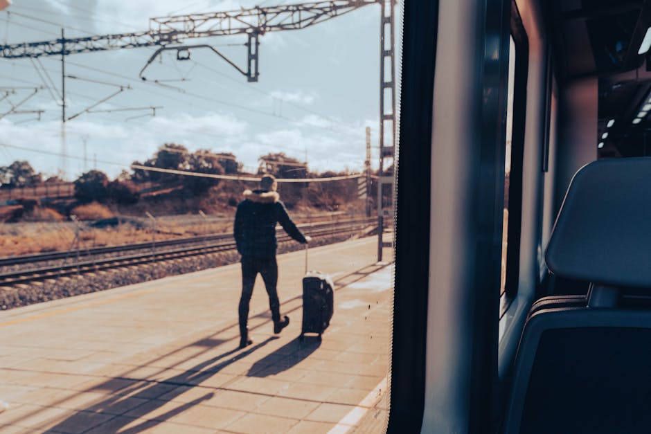 A person standing on a train platform outdoors, pulling a black wheeled suitcase with a telescopic handle. The individual is dressed in dark clothing and faces away from the camera, looking towards the railway tracks and distant trees and structures under a partly cloudy sky. The platform features a yellow safety line along the edge, with overhead electrical wires visible above. Inside the train, the right side of the image shows a dark blue car seat and part of the window frame, indicating the photo is taken from within the vehicle. This scene relates to home relocation or travel preparation, typical of moving or transport services provided by Man with Van Carshalton, during furniture transport or packing and moving logistics.
