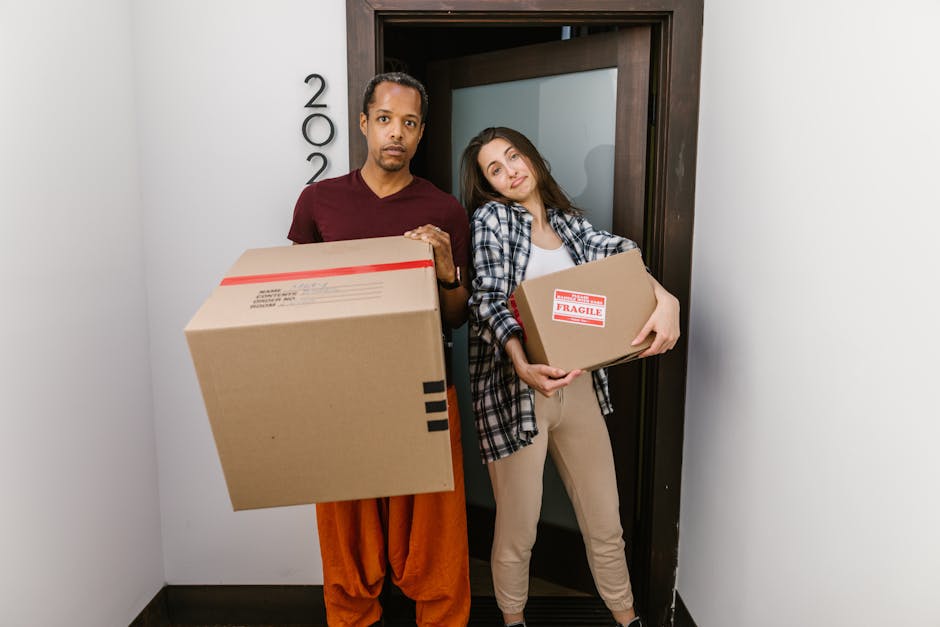 A man and a woman stand inside a doorway of a residential property, each holding a cardboard box during a home relocation process. The man, dressed in orange trousers and a maroon t-shirt, has a large cardboard box with black tape on the sides. The woman, wearing beige trousers and a plaid shirt, holds a smaller cardboard box labeled 'Fragile.' They appear to be preparing for or in the midst of loading or unloading items as part of a moving service. Behind them, the doorway features a dark brown door frame and a frosted glass panel. The scene is lit naturally, illustrating typical packing and furniture transport activities involved in house removals, often coordinated by services like Man with Van Carshalton. This image reflects the careful handling and organized packing associated with efficient home relocation, fitting within a context of professional removals near Honeywood Museum in Carshalton.