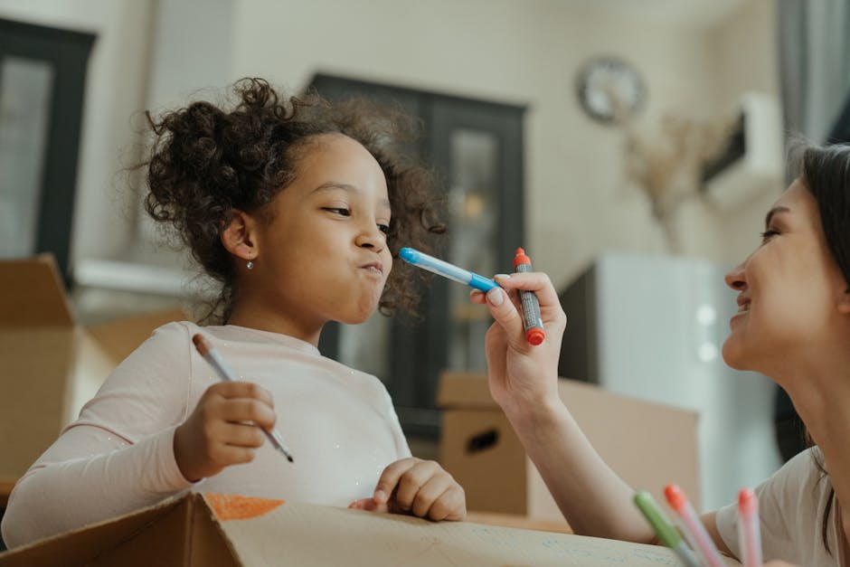 Inside a home during a packing and moving process, a woman is using a red marker to draw on a brown cardboard moving box, while a young girl with curly hair, wearing a light pink top, holds a thin black pen and stands nearby. The background features a dark wooden cabinet with glass doors, a wall clock, and a light-colored wall with a mounted skull or decorative object. Natural light illuminates the room, highlighting the activities related to home relocation, packing, and preparing items for transportation. The scene reflects the steps involved in furniture transport and packing for a house move, with moving boxes and packaging materials visible, supporting the ongoing process of local house removals, as provided by Man with Van Carshalton.