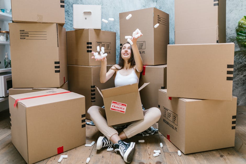 A man and a woman stand inside a doorway of a residential property, each holding a cardboard box during a home relocation process. The man, dressed in orange trousers and a maroon t-shirt, has a large cardboard box with black tape on the sides. The woman, wearing beige trousers and a plaid shirt, holds a smaller cardboard box labeled 'Fragile.' They appear to be preparing for or in the midst of loading or unloading items as part of a moving service. Behind them, the doorway features a dark brown door frame and a frosted glass panel. The scene is lit naturally, illustrating typical packing and furniture transport activities involved in house removals, often coordinated by services like Man with Van Carshalton. This image reflects the careful handling and organized packing associated with efficient home relocation, fitting within a context of professional removals near Honeywood Museum in Carshalton.