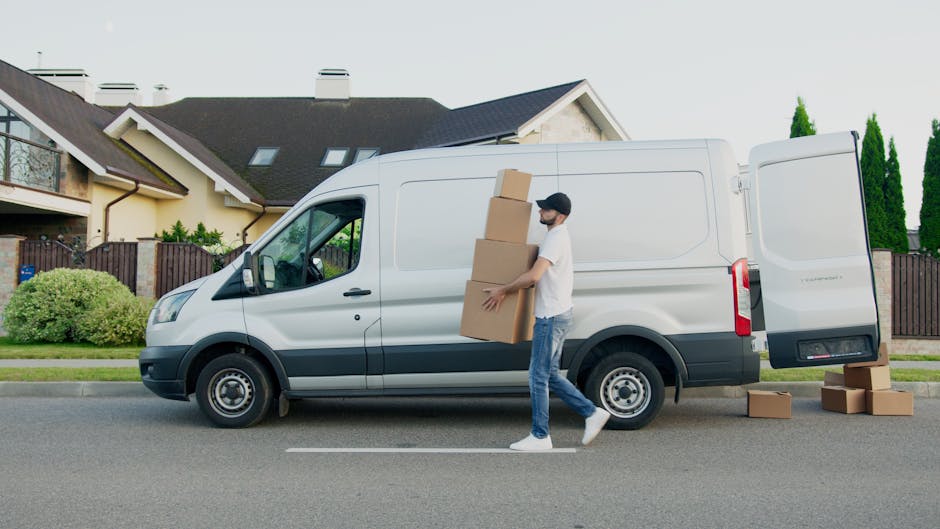 A man in casual clothing, including a white t-shirt, jeans, and a dark cap, is seen carrying three stacked cardboard boxes while walking beside a white commercial van parked on a residential street. T