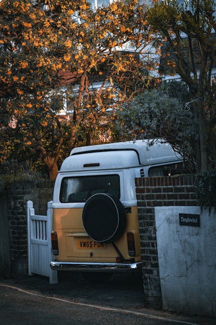 A white and yellow vintage van parked partially behind a brick wall and white gate in a residential area, with trees and foliage around, illuminated by warm evening light. The van appears ready for home relocation or furniture transport, with the rear door closed and a spare tire mounted on the back. The scene is part of a moving process, showcasing loading or unloading in a typical street environment. This image reflects the logistical aspects of packing and moving, relevant to house removals services provided by Man with Van Carshalton, particularly in areas like Carshalton with easy van access for efficient transport.