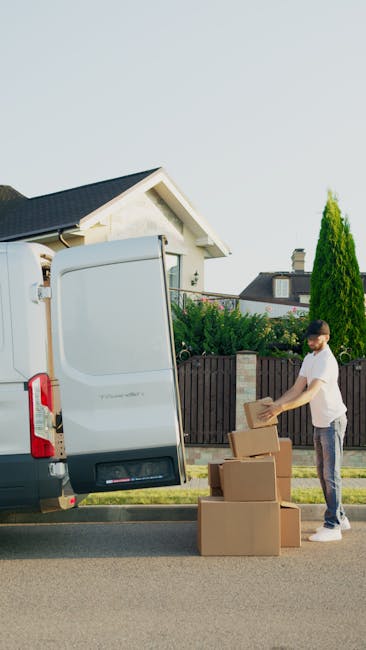 A man wearing a white t-shirt and dark jeans is standing outdoors beside a white moving van with its back doors open. He is handling cardboard boxes, some stacked on the pavement and others being loaded into the van for a home relocation. The boxes are made of brown cardboard, with one partially wrapped in plastic film for protection. Inside the van, visible packing materials include a plastic sheet and bubble wrap. The environment features a suburban street setting with a wooden fence, green shrubbery, and a house with a pitched roof and chimney in the background, under a clear blue sky. The scene captures the process of packing and loading for furniture transport and moving, with the focus on careful handling of household items using moving equipment such as trolleys or blankets, indicative of professional removals by Man with Van Carshalton.