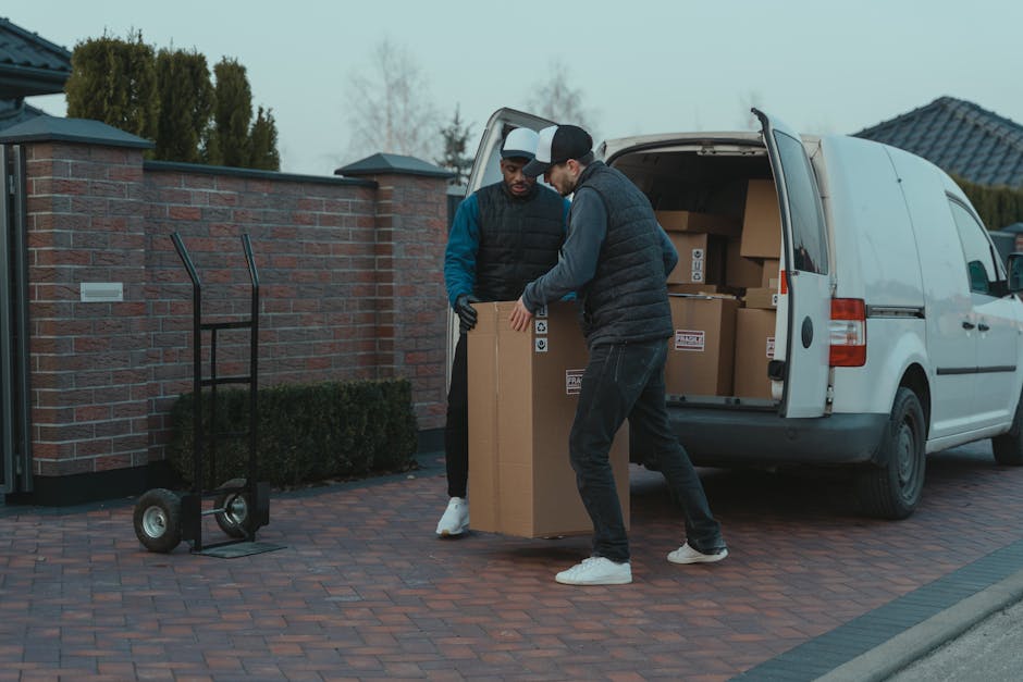 A white and yellow vintage van parked partially behind a brick wall and white gate in a residential area, with trees and foliage around, illuminated by warm evening light. The van appears ready for home relocation or furniture transport, with the rear door closed and a spare tire mounted on the back. The scene is part of a moving process, showcasing loading or unloading in a typical street environment. This image reflects the logistical aspects of packing and moving, relevant to house removals services provided by Man with Van Carshalton, particularly in areas like Carshalton with easy van access for efficient transport.