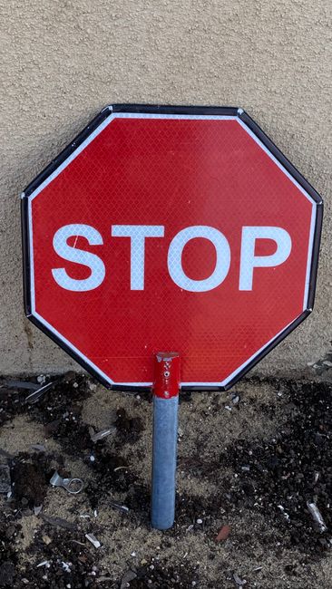 An image showing a red and white stop sign attached to a stone wall at a street corner, with the sign facing outward for oncoming traffic. Next to it, mounted on a plain white wall, is a circular no p