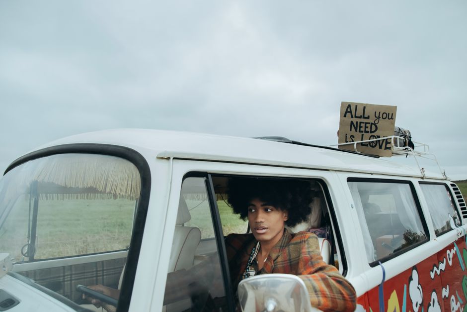 A young woman with natural curly hair sitting inside the front cabin of a vintage white van decorated with colourful, illustrative patterns on its side panels, during daytime with cloudy skies. She is wearing a plaid shirt and appears to be looking outside, possibly overseeing a home relocation process. A cardboard sign with the message 'ALL you NEED is LOVE' is attached to the roof of the van, secured with ropes. The van is parked on an open grassy area, with the background showing a field and an overcast sky. The scene captures aspects of furniture transport and packing during a move, with the woman involved in a loading or unloading activity, supported by the context of professional removals by Man with Van Carshalton, illustrating the moving process related to house removals and transport logistics.