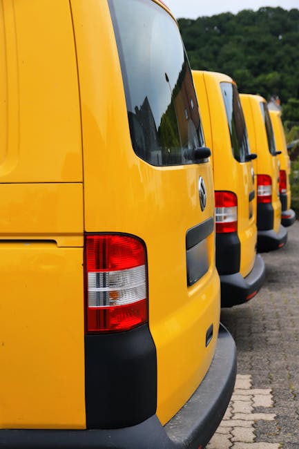 Four yellow commercial vans are parked in a row on a paved surface, with their rear ends facing the camera. The vans have black bumpers and red and white tail lights. The sky overhead is partly cloudy, and a hillside covered in greenery is visible in the background. These vehicles are likely used for home relocation or furniture transport services, and the image may relate to planning or executing a house removal. The setting suggests a loading area where packing materials or furniture are being prepared for transportation, consistent with professional removals by Man with Van Carshalton. No interior details or objects are visible inside the vans, and the focus is on the exterior parked vehicles, representing logistical aspects of moving services.