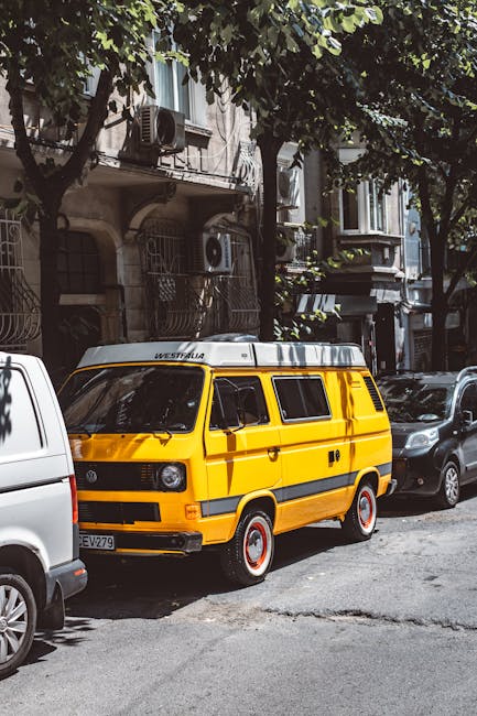A yellow Volkswagen Westfalia campervan is parked on the side of a narrow urban street during daylight hours, with trees providing partial shade. The van is positioned front-on, showing its distinctive boxy shape, black window frames, and white roof with the Westfalia logo. Surrounding the campervan are other parked vehicles, including a silver van and a dark-colored car, all situated on a paved street with typical residential buildings having balconies, air conditioning units, and window grills in the background. The area appears calm and suitable for home relocation or furniture transport, consistent with services offered by Man with Van Carshalton, who may sometimes use such vehicles for moving and packing logistics.