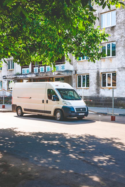 A white commercial van belonging to Man with Van Carshalton is parked on a street in front of a multi-storey residential building with concrete walls and multiple windows. The van is positioned parallel to the pavement, with the front facing to the right of the image. Overhanging branches from a large leafy tree cast shadows on the road and the van. The pavement beside the van is clear, and there are red and white bollards along the curb, indicating a designated parking or loading zone. Inside the open doorway of the building, visible are several cardboard boxes and packing materials, suggesting a home relocation or furniture transport process managed by [COMPANY_NAME]. The building has a weathered appearance, with some visible stains on the concrete exterior and decorative snowflake motifs on the balcony railing. The scene is lit by daylight, emphasizing the urban environment and the logistical aspects of house removals and moving services that [COMPANY_NAME] provides through their vehicle and loading area at this site.