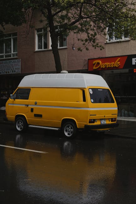 A yellow Volkswagen Westfalia campervan is parked on the side of a narrow urban street during daylight hours, with trees providing partial shade. The van is positioned front-on, showing its distinctive boxy shape, black window frames, and white roof with the Westfalia logo. Surrounding the campervan are other parked vehicles, including a silver van and a dark-colored car, all situated on a paved street with typical residential buildings having balconies, air conditioning units, and window grills in the background. The area appears calm and suitable for home relocation or furniture transport, consistent with services offered by Man with Van Carshalton, who may sometimes use such vehicles for moving and packing logistics.